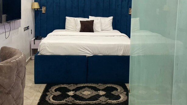 Ein modernes Schlafzimmer mit blauem Kopfteil, einem flachbildfernseher an der Wand, einem Nachttisch mit Lampe und einer Glastrennwand.