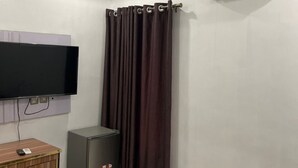Ein Zimmer mit Flachbildfernseher an der Wand, einem kleinen Kühlschrank, einem Schrank mit Fernbedienung, einem Vorhang und einer Klimaanlage.