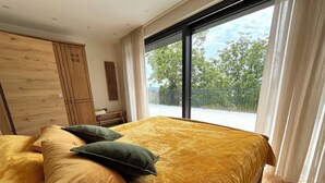 A bedroom with a large bed, wooden wardrobe, and a view of the outdoors through a large window.
