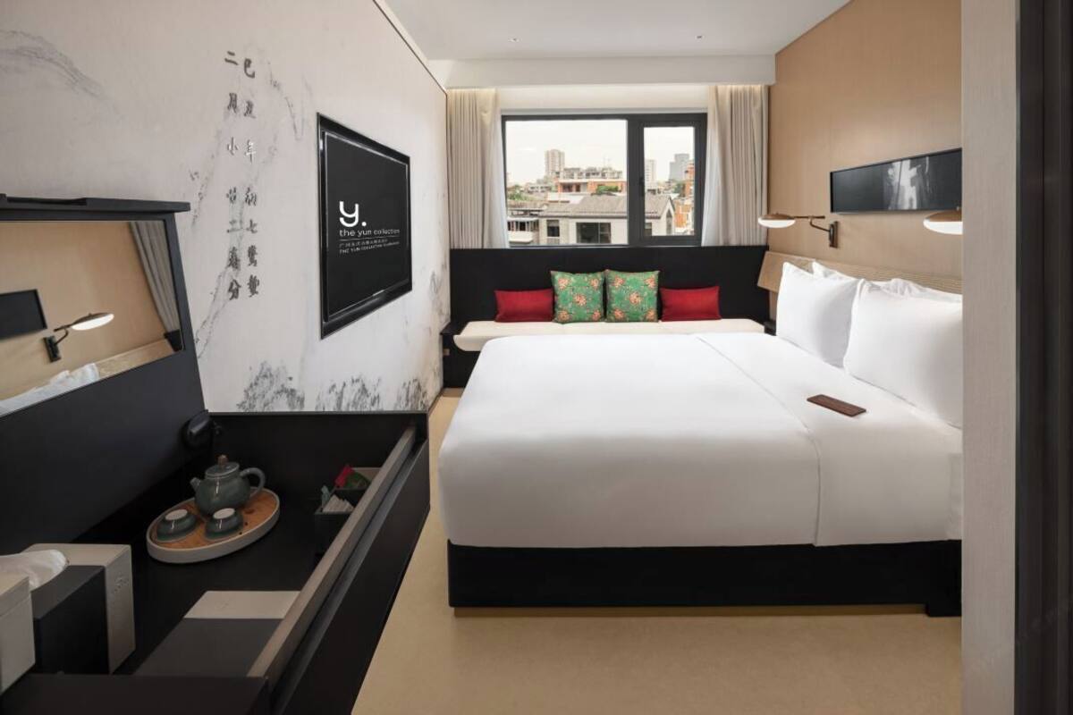 A hotel room with a large bed, a television, a desk with a teapot, and a view of the city through the window.
