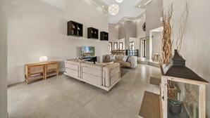 Un salon spacieux avec un grand canapé beige, une console en bois et un téléviseur. On y trouve des luminaires au plafond et une plante décorative.