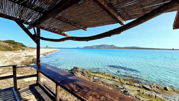 Een houten terras met rieten dak met uitzicht op een strand en helderblauw water.