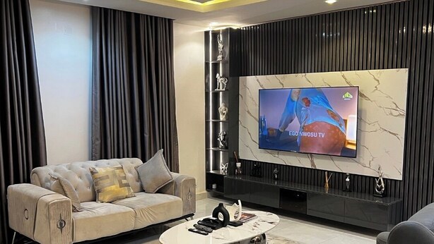 Een moderne woonkamer met een grote flatscreen televisie, een gestoffeerde bank, een salontafel en een donkerblauwe fauteuil.