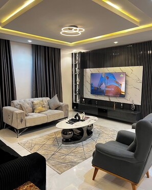 A modern living room with a large flat-screen TV, a tufted sofa, a coffee table, and a dark armchair. - Theodora Apartment (Abuja)