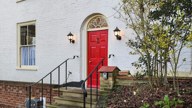 A white building with a red door, a window with a blue curtain, and a small garden area with steps leading to the door.