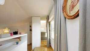 Ein schmaler Flur mit Holzboden, weißen Wänden und einer Tür zu einem anderen Raum. Regale mit Dekorationselementen und ein gerahmtes Bild sind an der Wand angebracht.