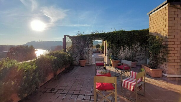 Eine Dachterrasse mit Blick auf einen Fluss und ferne Hügel, mit Topfpflanzen und zwei Holzstühlen mit roten Kissen.