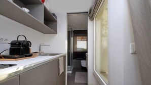 Ein enger Flur führt zu einem Badezimmer mit Toilette und Waschbecken. Rechts befindet sich ein Fenster, links ein Küchenbereich mit Wasserkocher und Toaster.