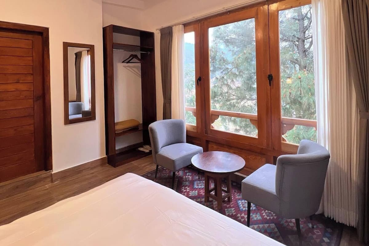 A hotel room with a bed, two armchairs, a small table, a wardrobe, and a window with a view of trees.