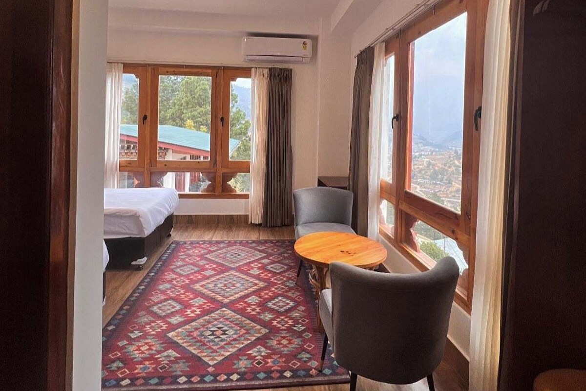 A room with a bed, a small table, a chair, and large windows offering a view of the outside.