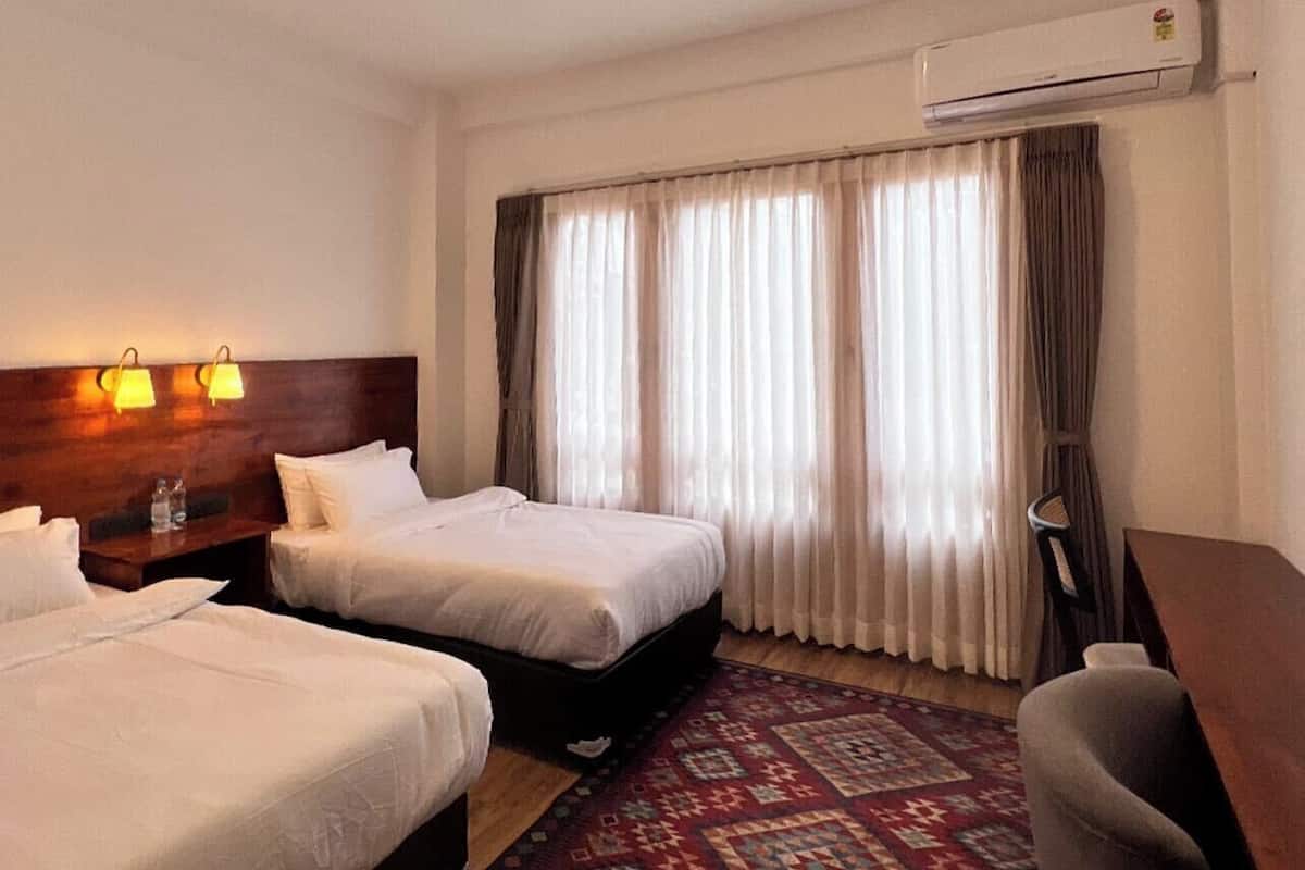A hotel room with two beds, a desk, a chair, a patterned rug, and an air conditioning unit.