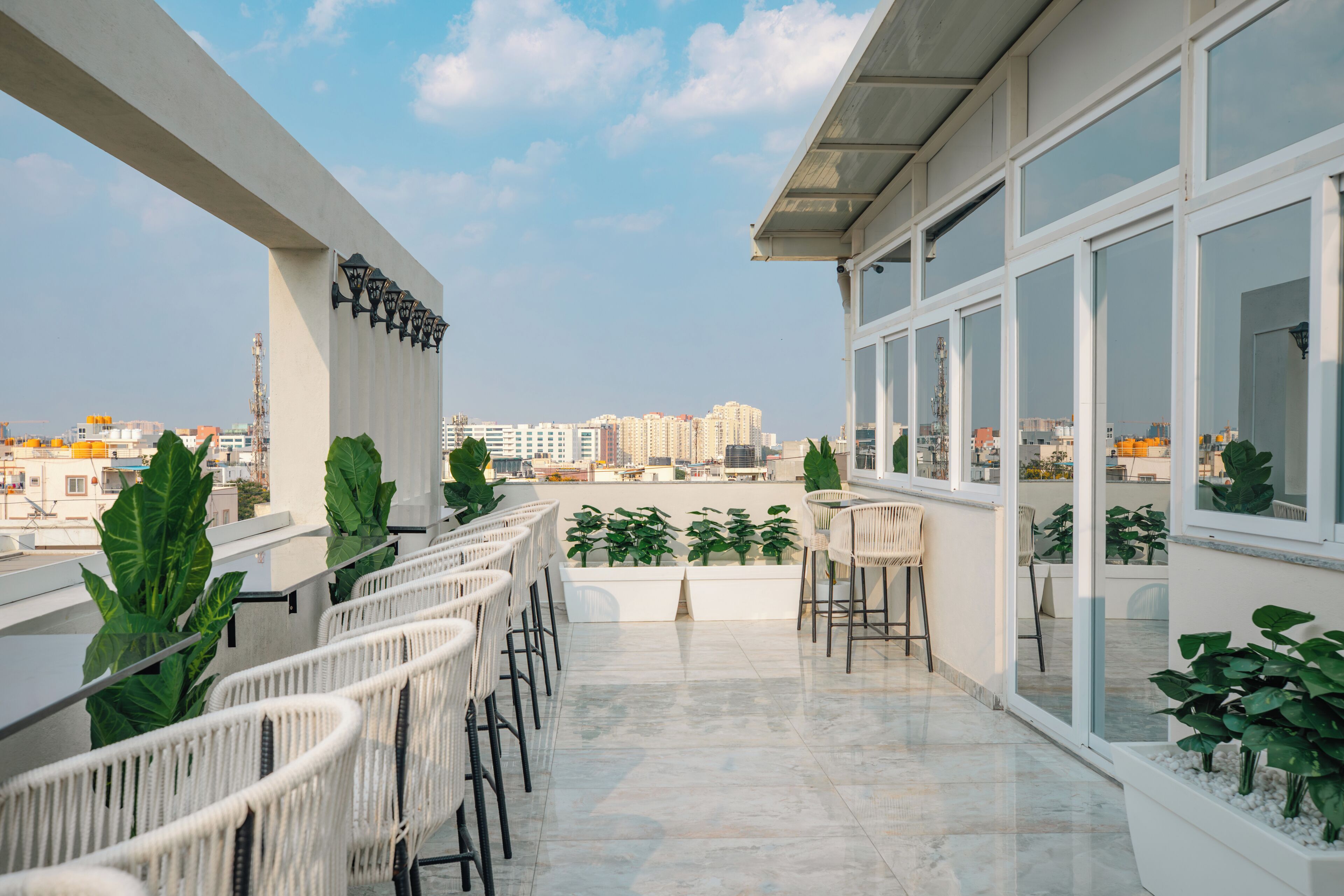 A rooftop terrace with white wicker chairs, potted plants, and a city view.