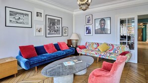 Uma sala de estar com um sofá azul, um sofá colorido, uma mesa de centro redonda e uma cadeira vermelha.