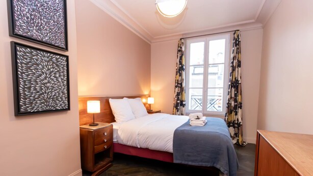 Ein Schlafzimmer mit Bett, Nachttischen, einem Schminktisch, einem Fenster mit Vorhängen und Kunstwerken an der Wand.