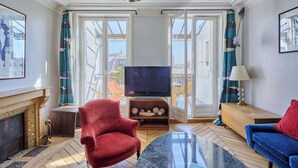 Uma sala de estar com lareira, uma poltrona vermelha, um sofá azul, uma televisão sobre um suporte de madeira e vista para edifícios através de grandes portas de vidro.