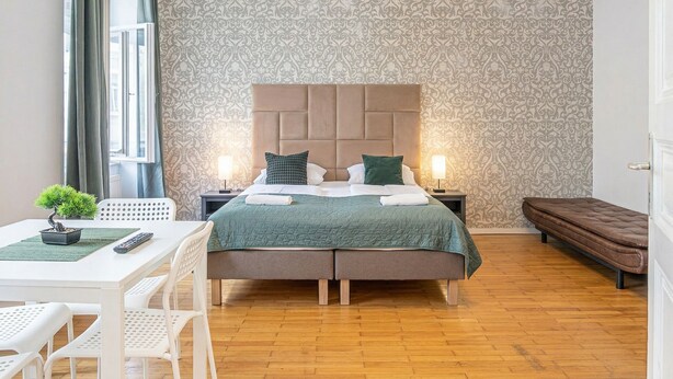 A bedroom with a patterned wallpaper, a bed with a headboard, bedside tables, a bench, a dining area with a table and chairs, and a small plant. - Lovely 2BR Near Belvedere Palace (Vienna)
