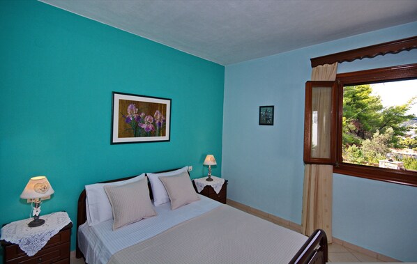 A bedroom with teal walls, a bed with white bedding, two bedside tables with lamps, and a window with a view of greenery. - Kamelia Villas in Alonissos (Alonissos)