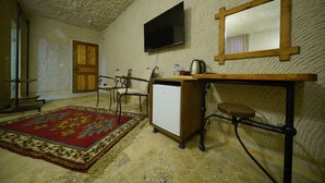 A room with a stone floor, a wooden door, a television mounted on the wall, a small refrigerator, a wooden bench, and a patterned rug. - nirvana cave house hotel (Nevsehir)