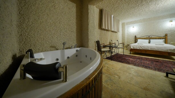 A bathroom with a freestanding bathtub, a sink, and a chair. There is a bed with a wooden headboard in the adjacent room. - nirvana cave house hotel (Nevsehir)