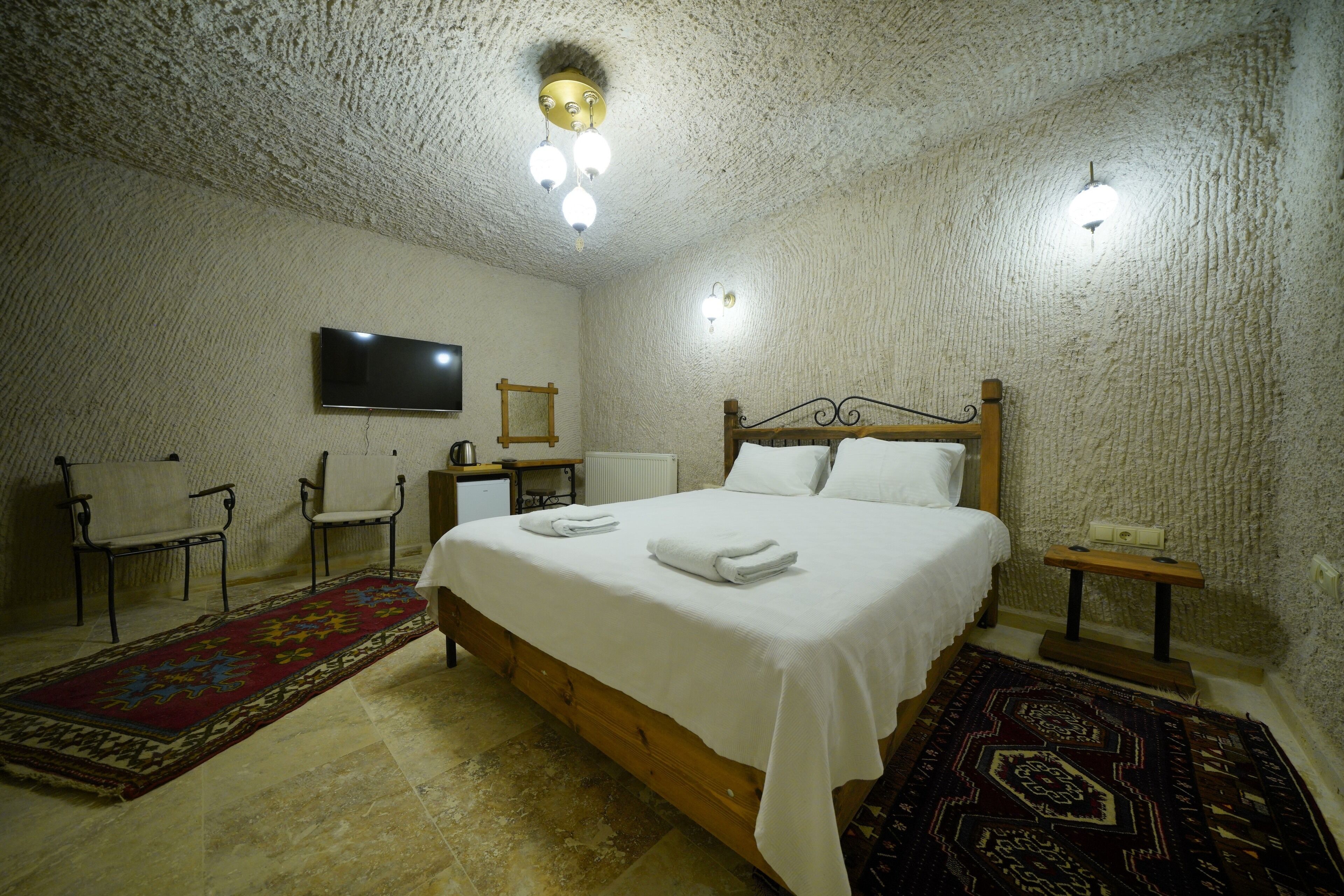 A bedroom with a wooden bed, white linens, a red and blue patterned rug, a small bedside table, and a wall-mounted TV.