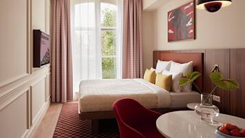 A hotel room with a bed, a television, a window with curtains, a painting on the wall, and a table with a bowl of fruit.