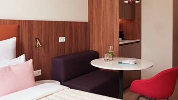 A hotel room with a bed, a round table, a red chair, a purple sofa, and a pendant light.