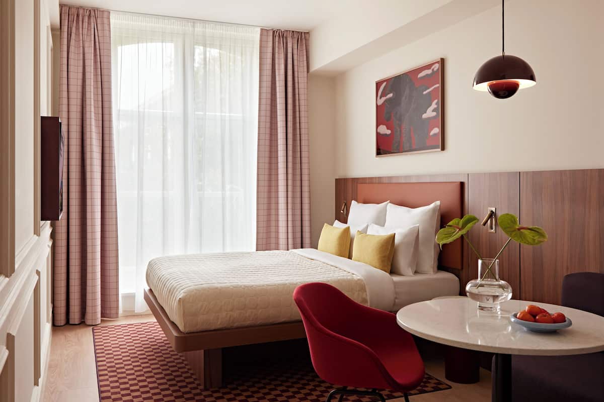 Hotelkamer met een bed, een rode stoel, een ronde tafel met een fruitschaal, een schilderij aan de muur en een raam met gordijnen.