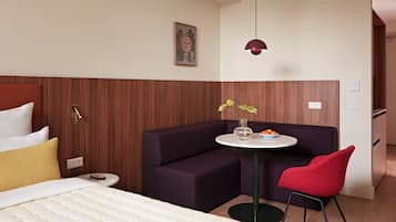 A hotel room with a bed, a sofa, a small table with a vase and oranges, a red chair, and a pendant light.
