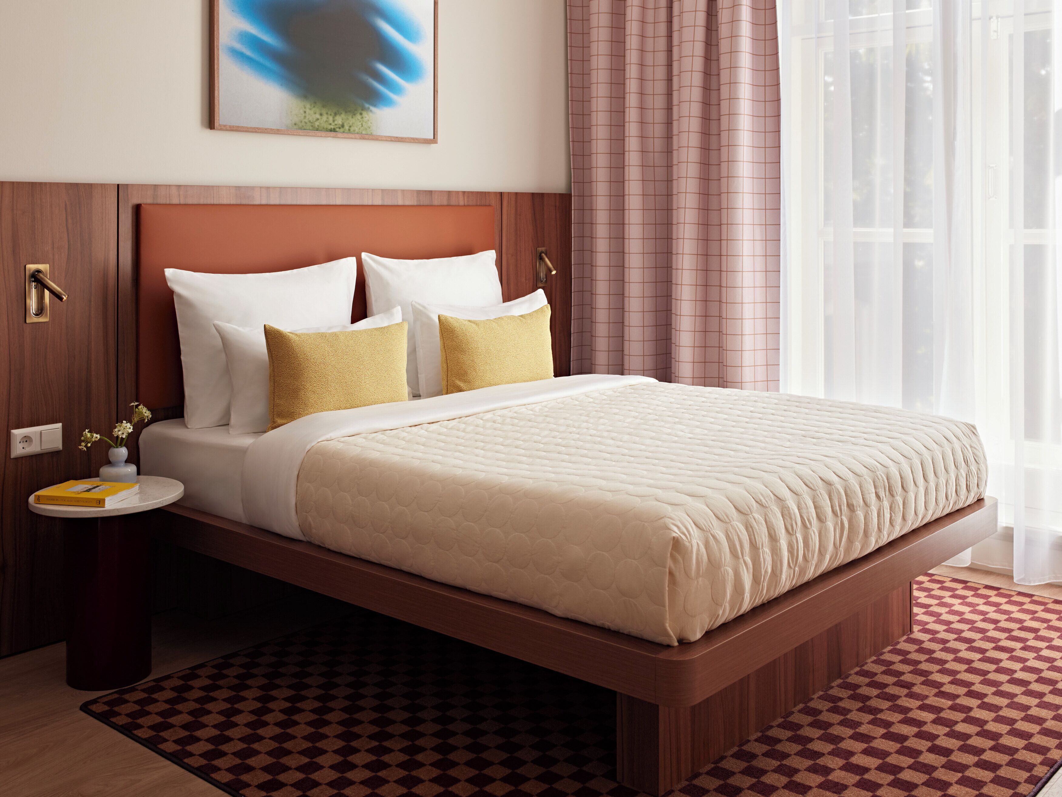 Habitación de hotel con cabecera de madera, cama grande con ropa de cama blanca y dorada, mesita de noche con un libro y un florero, y alfombra a cuadros.