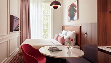 Een hotelkamer met een bed, een ronde tafel, een rode stoel, een paarse bank, een raam met gordijnen, een kunstwerk aan de muur en een welkomstbord.