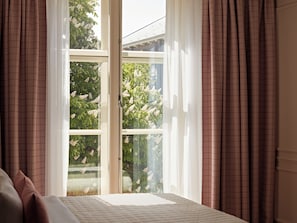 A room with a bed, a window with curtains, and a view of trees and a building outside.