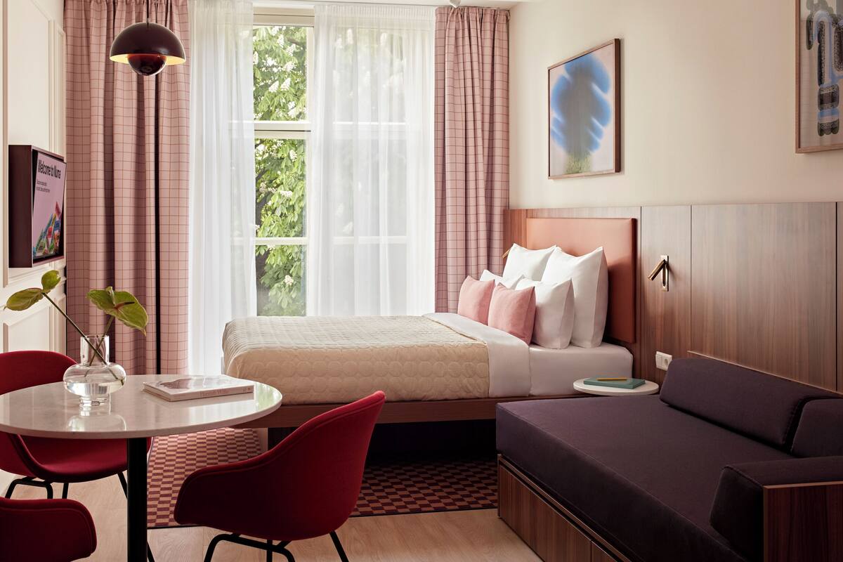 Een hotelkamer met een bed, een bank, een eettafel met stoelen, een hanglamp en een raam met gordijnen.