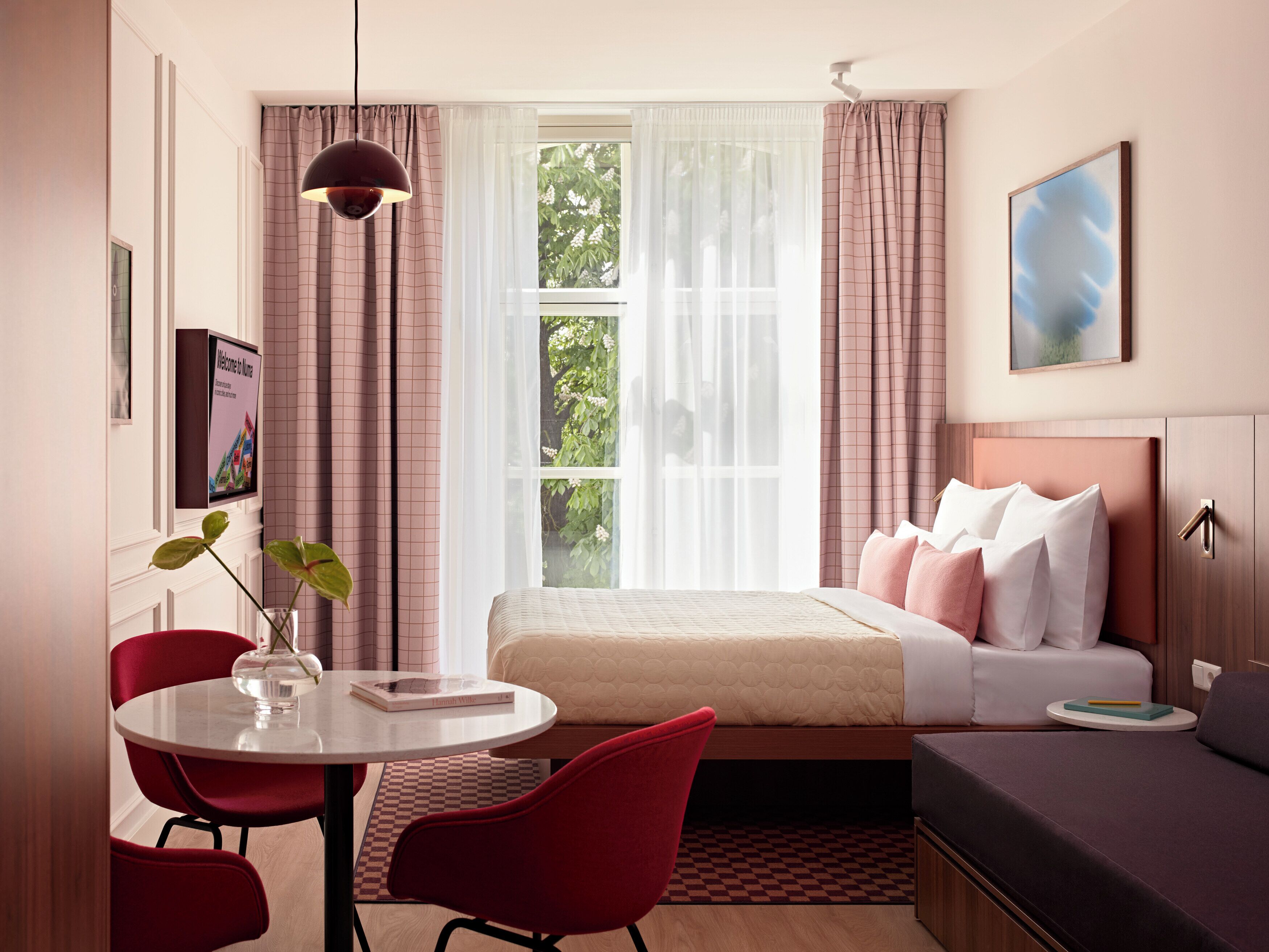 A hotel room with a bed, a desk with a chair, a window with curtains, a plant, and a painting on the wall.