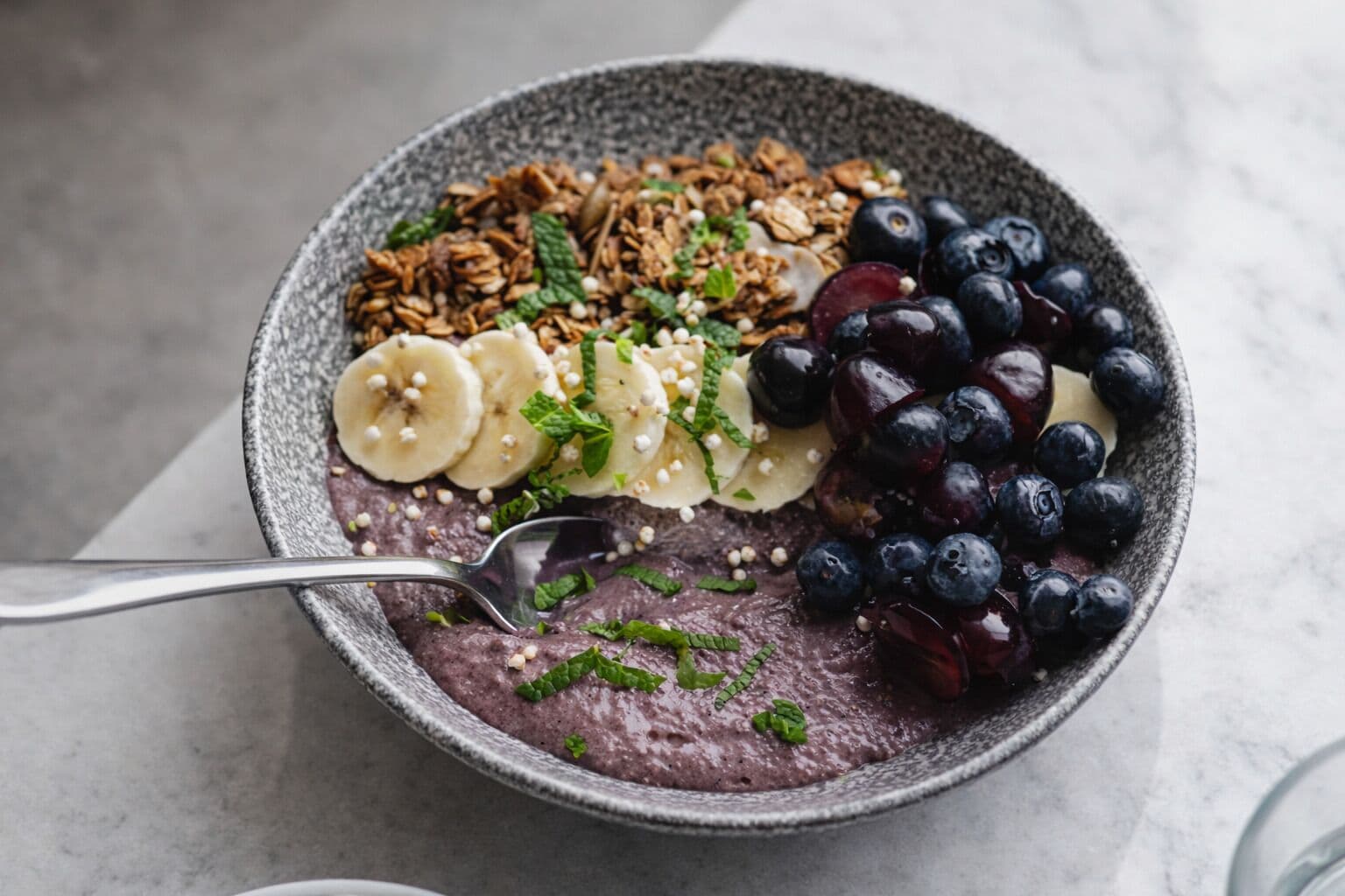 En skål med smoothie toppad med granola, bananskivor, blåbär och myntablad.