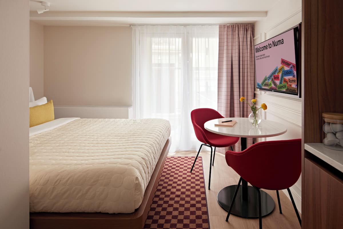 Een hotelkamer met een bed, een tafel met twee stoelen, een vaas met bloemen en een aan de muur bevestigde televisie waarop een welkomstboodschap staat.