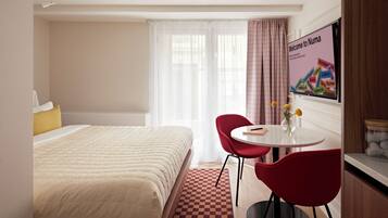 A hotel room with a bed, a table with two chairs, a vase with flowers, and a wall-mounted TV displaying a welcome message.