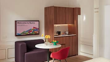 A modern hotel room with a dark purple sofa, a red chair, a white round table, and a kitchen area with wooden cabinetry.