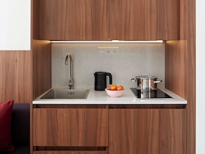 Eine kompakte Küche mit Spülbecken aus Edelstahl, einem schwarzen Wasserkocher, einem Topf auf einer Induktionskochplatte und einer Schüssel Orangen auf der Arbeitsplatte.