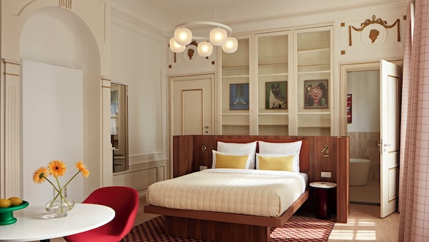Een slaapkamer met een bed, een rode stoel, een klein tafeltje met bloemen en een badkamer zichtbaar door een geopende deur.