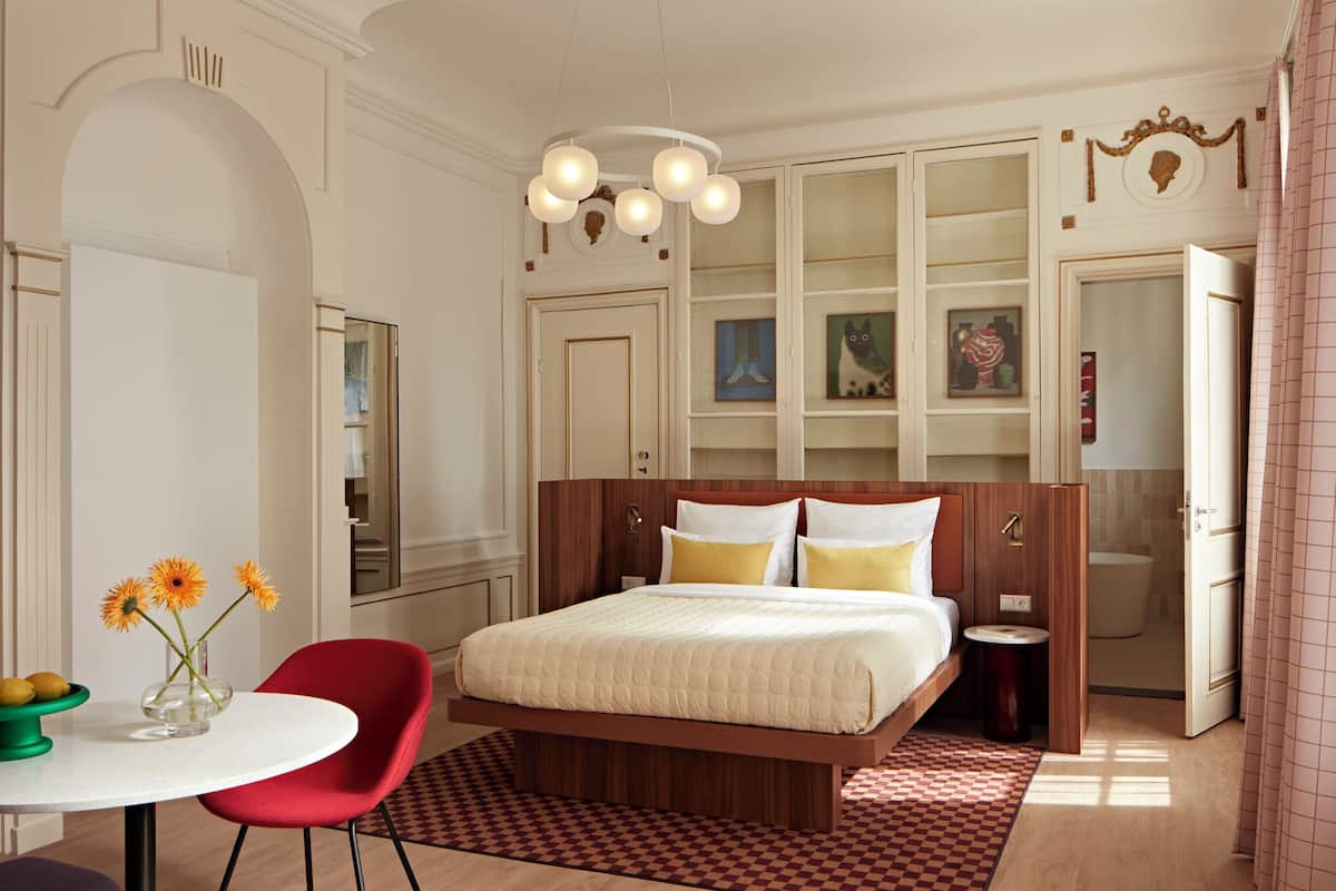Een slaapkamer met een bed, een rode stoel, een klein tafeltje met bloemen en een badkamer zichtbaar door een geopende deur.