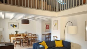 A modern living room with a blue sofa, wooden dining table, and chairs. There is a staircase in the background leading to an upper level. - Le Clos du Cornier - Peace & quiet & private pool (Barjac)