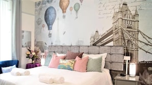 Ett hotellrum med en säng, kuddar, ett nattduksbord, en lampa och en väggmålning som föreställer en bro och varmluftsballonger.