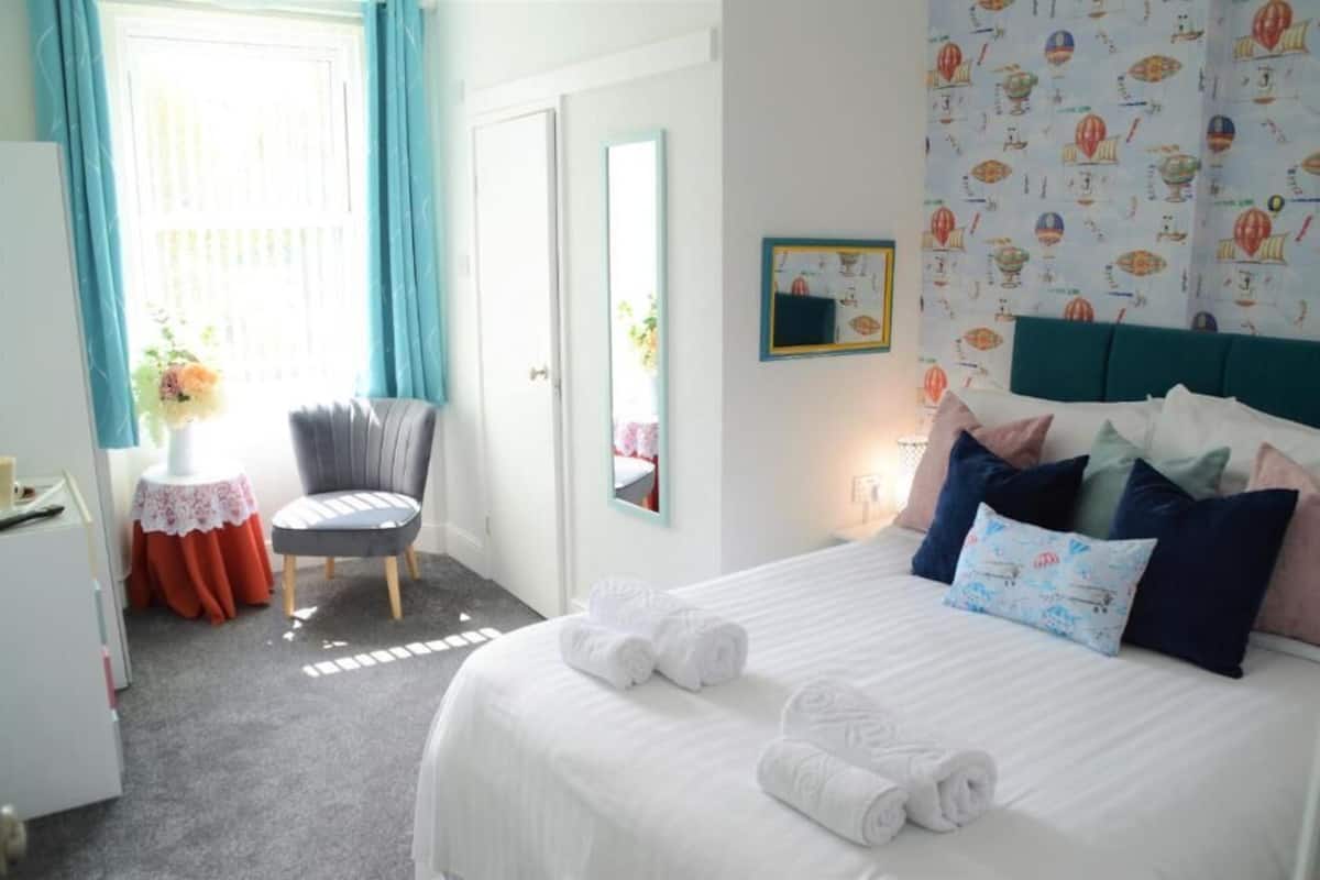 A hotel room with a bed, pillows, a chair, a small table with a vase, and a window with curtains.