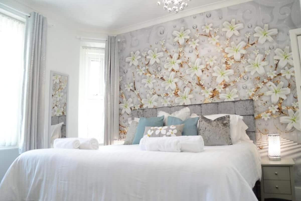 A bedroom with a large bed, a headboard with a floral design, a bedside table with a lamp, and a window with curtains.