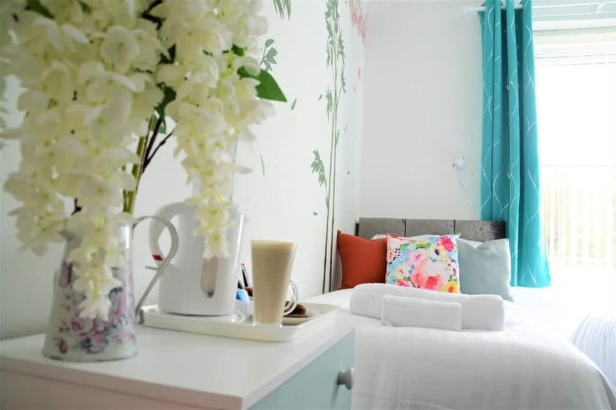 A neatly arranged bedroom with a bed, pillows, a bedside table with a coffee maker and a vase of flowers, and a window with curtains.