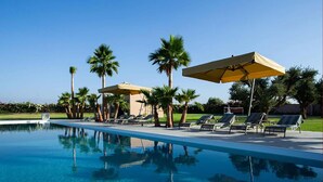 Zona de piscina amb gandules i ombrel·les, envoltada de palmeres i un cel serè.