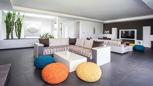 Ein modernes Wohnzimmer mit einem Ecksofa, runden Hocker, einem Flachbildfernseher und einem großen Fenster mit Pflanzen.
