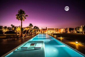 Un espace piscine éclairé la nuit, avec des chaises longues et la lune dans le ciel.