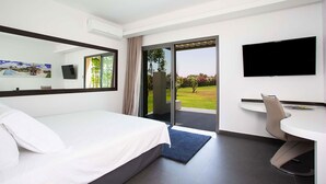 Habitació d'hotel moderna amb un llit, una taula de treball, una cadira, un televisor i una porta corredissa de vidre que dona a un balcó amb vista a un paisatge verd.