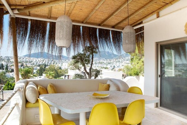 Un área de estar al aire libre techada, con una mesa y sillas amarillas, con vistas a un paisaje urbano y una montaña al fondo.